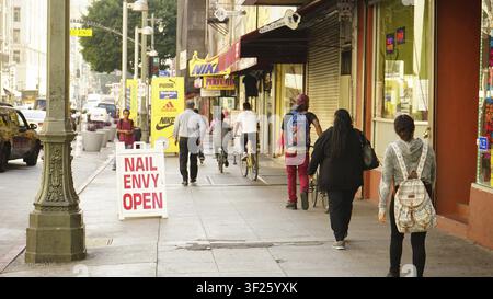 Fußgänger gehen entlang eines Stadtrandsteins, flankiert von verschiedenen Geschäften und Straßenschildern, DTLA -- Los Angeles, Kalifornien, USA Stockfoto