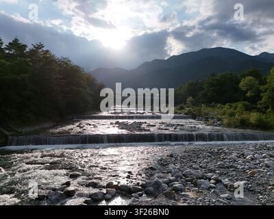 Sonnenlicht bricht durch die Wolken über einem fließenden Fluss. Felsen und Wasser bilden sanfte Kaskaden über den Bach. Üppige grüne Bäume säumen beide Ufer unter einem Stockfoto