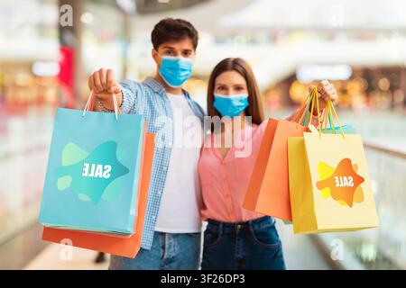 Junges Paar lächelt mit Einkaufstüten in der Mall, trägt Schutzmasken, Verkaufsbegriff Stockfoto