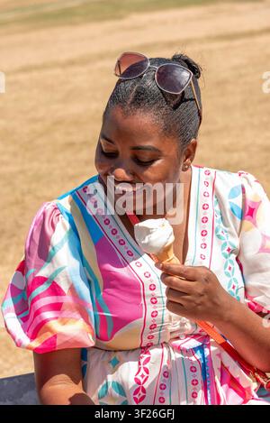 Nottingham England UK. 11.07.2026. Eine lächelnde Frau genießt an einem warmen Tag eine kalte Leckerei, trägt ein gemustertes Kleid und eine Sonnenbrille auf dem Kopf. Stockfoto