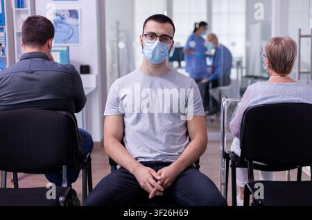 Lächelnder junger Patient sitzt auf einem Klinikstuhl im Wartezimmer, während ein Arzt im Hintergrund arbeitet und eine Assistentin am Computer tippt. Stockfoto