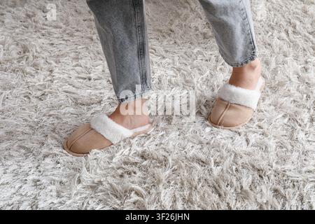 Person, die bequeme beige Pantoffeln mit weißer Pelzfütterung auf einem weichen, zotteligen weißen Teppich zu Hause trägt Stockfoto