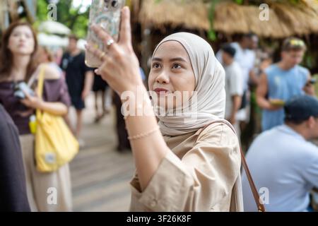 Asiatisch-indonesische Frau nutzt ein Smartphone, um Fotos zu machen, während sie einen belebten Markt auf Bali, Indonesien, erkundet. Reise-, Technologie- und Lebensstilkonzept. Stockfoto