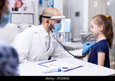 Kinderarzt mit einer Gesichtsmaske hört das Herz eines Kindes mit einem Stethoskop während einer medizinischen Konsultation in einer Praxis, verbindet Konsultation und Gesundheitsversorgung in einer klaren Klinik. Stockfoto