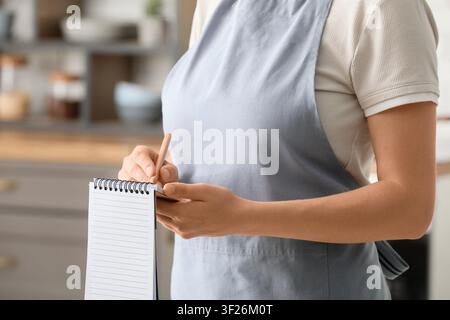 Frau in einer hellblauen Schürze, die in einer Küchenumgebung mit einem Bleistift in ein Spiralnotizbuch schreibt Stockfoto