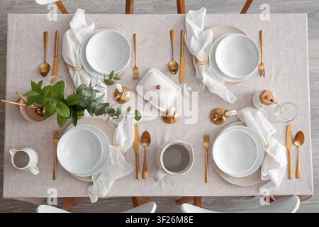Eleganter Esstisch mit weißen Tellern, goldenem Besteck und grünen Eukalyptuszweigen für ein festliches Mahl Stockfoto