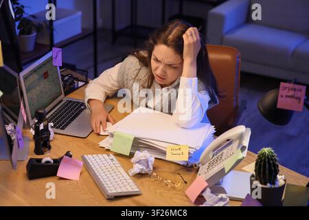 Überwältigte junge Frau, die spät in der Nacht in einem schwach beleuchteten Büro arbeitet und mit einer schweren Arbeitslast und knappen Fristen kämpft Stockfoto