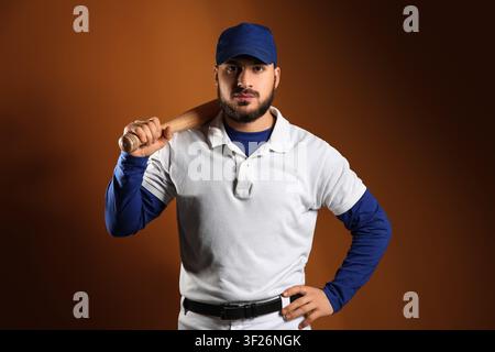 Junger bärtiger Baseballspieler mit blauer Mütze und weißer Uniform, der einen Holzschläger über der Schulter hält, der Athlet steht selbstbewusst vor einem braunen Hintergrund Stockfoto