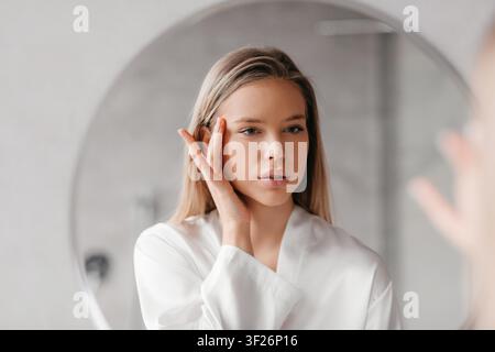 Junge Frau, die vor dem Spiegel zu Hause eine Hautpflegecreme aufträgt, Selbstpflege-Konzept Stockfoto