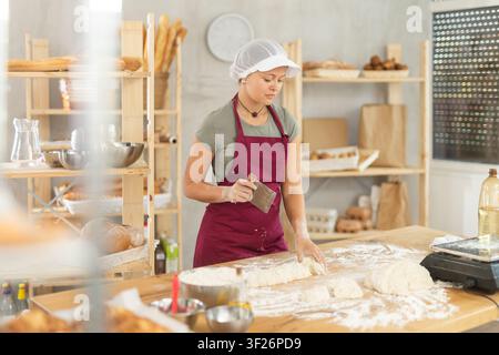Bäckerin schneidet rohen Teig mit einem Messer und wiegt Stücke rohen Teigs auf einer Waage für die Herstellung von Croissants Stockfoto