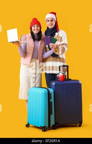 Nettes junges Paar mit Laptop, Pässen und Koffern auf gelbem Hintergrund. Winterurlaubskonzept Stockfoto