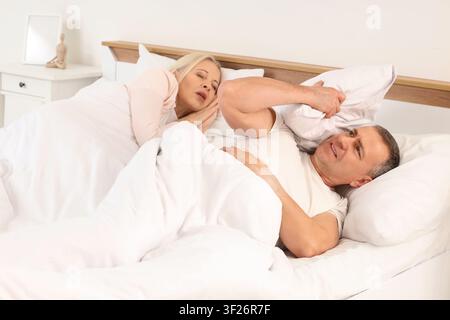 Gereizter reifer Mann, der mit seiner schnarchenden Frau im Schlafzimmer liegt Stockfoto