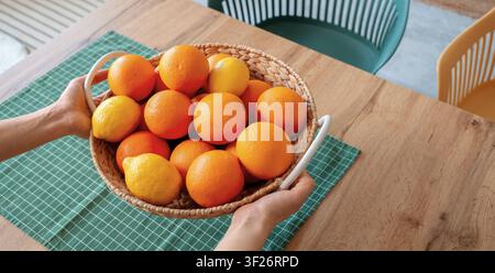 Hände halten einen geflochtenen Korb gefüllt mit frischen Orangen und Zitronen auf einem Holztisch mit einem grün karierten Tuch, der Korb ruht auf einem warmen Holztisch Stockfoto