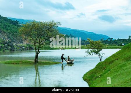 Zwei Personen stehen auf einem kleinen Boot nahe dem Ufer eines ruhigen Sees, umgeben von grünem Gras und Bäumen mit einer Berglandschaft im Hintergrund. Stockfoto