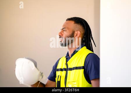 Profilansicht eines afroamerikanischen Ingenieurs mit erschöpftem Blick, der einen weißen Schutzhelm in einem unfertigen Wohngebäude am Tag hält, Gefühl von Stockfoto