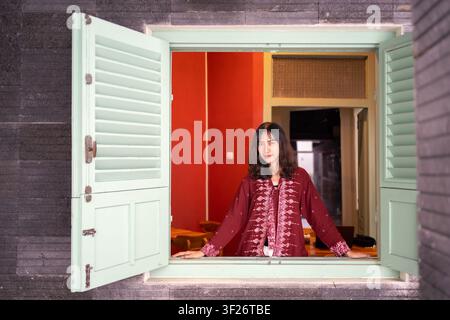 Porträt einer jungen indonesischen Frau in Batik-Mode, die an einem offenen Fenster steht. Konzept von Kultur, Lebensstil und zeitgenössischer Identität. Stockfoto