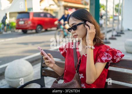 Stilvolle indonesische Frau trägt eine Batikbluse und eine Sonnenbrille und überprüft ihr Smartphone auf einer öffentlichen Bank. Konzept der modernen Kommunikation. Stockfoto