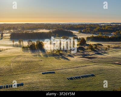 Luftaufnahme einer ländlichen Landschaft mit Morgennebel, Heuballen auf den Feldern und Bäumen während der Goldenen Stunde in Vidzeme, Lettland. Stockfoto
