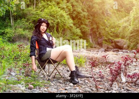 Eine junge asiatische Frau sitzt auf einem Campingstuhl in einer Waldumgebung und genießt ein heißes Getränk und die friedliche Landschaft Stockfoto