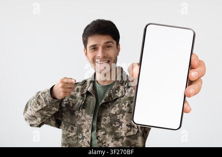 Lächelnder junger Mann in Militäruniform, der ein Smartphone mit leerem Bildschirm hält, Mockup Stockfoto
