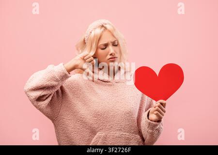 Traurige junge Frau, die ein Herzsymbol ablehnt, rosa Hintergrund, emotionaler Ausdruck, Liebeskonzept Stockfoto