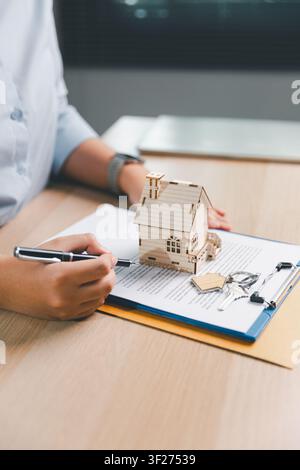 Szenen der Unterzeichnung eines Immobilienvertrags mit Holz-Hausmodell, Stift, Schlüsseln und Dokumenten auf dem Bürotisch, die den Immobilienkauf, Hypothekenunterlagen, m zeigen Stockfoto