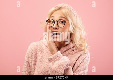 Überraschte junge Frau mit Brille, die ein Geheimnis flüstert, rosa Hintergrund Stockfoto