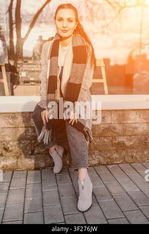 Damenschal Mode Junge Frau mit kariertem Schal und Mantel sitzt während der goldenen Stunde des Sonnenuntergangs auf einer Steinmauer Stockfoto