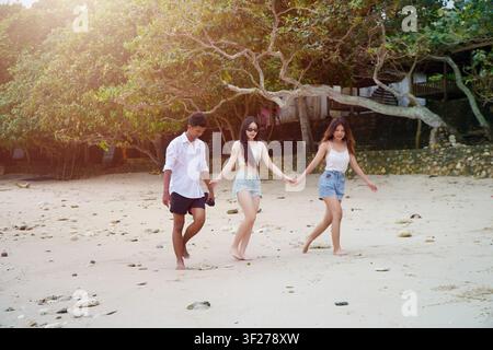 Drei junge asiatische Freunde laufen barfuß an einem tropischen Strand, halten sich an den Händen und genießen einen sonnigen Tag am Meer. Stockfoto
