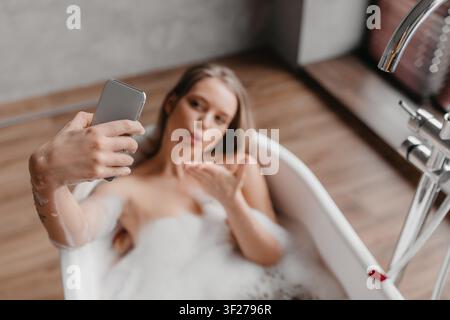 Junge Frau sitzt in der Badewanne und macht mit dem Smartphone ein Selfie, Entspannungskonzept Stockfoto