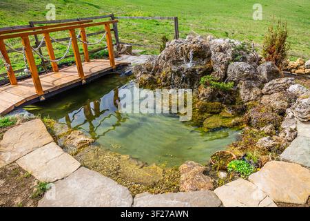 Hölzerne Fußgängerbrücke über einen ruhigen Gartenteich mit einem kleinen Wasserfall Stockfoto