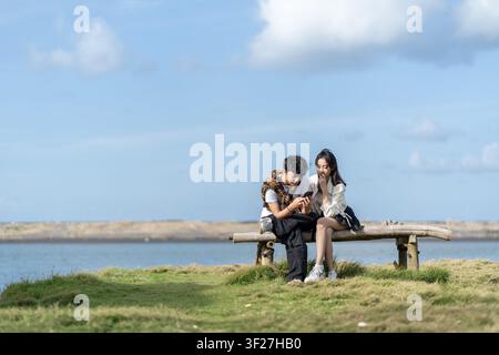 Lächelndes junges asiatisches indonesisches Paar sitzt auf einer Holzbank am Wasser und teilt ein Smartphone im Freien. Lässiger Lebensstil, moderne Beziehung, ein Stockfoto