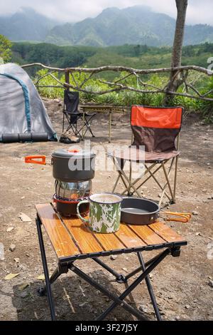 Camping-Tasse, tragbarer Herd und Kochgeschirr auf einem Tisch mit Zelt und Berglandschaft, die Outdoor-Kochen und Abenteuerreisen repräsentieren. Stockfoto