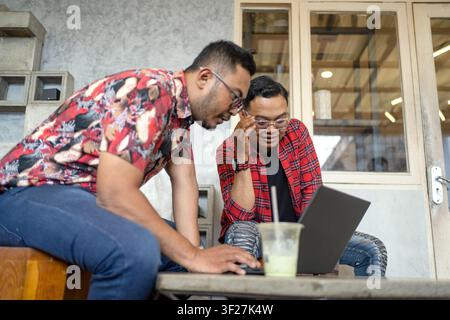 Zwei indonesische Unternehmer, die während eines lockeren Geschäftsmeetings in einem modernen Café Ideen diskutieren und Informationen auf einem Laptop durchsehen. Stockfoto