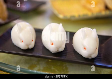 Drei entzückende, durchscheinende weiße, kaninchenförmige Gelee-Desserts mit kleinen roten Augen, die in einer Reihe auf einer langen schwarzen Servierschale angeordnet sind. Stockfoto