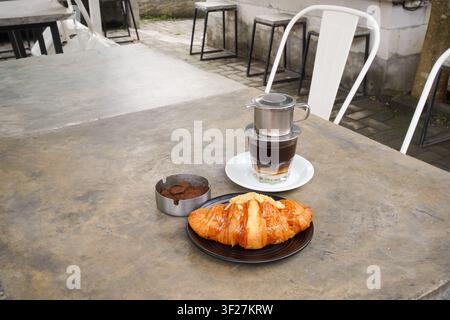 Frisches Mandelkroissant serviert mit traditionellem vietnamesischem Filterkaffee an einem gemütlichen Außentisch eines Cafés. Morgenfrühstücksambiente mit rustikaler Note und na Stockfoto