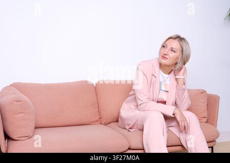 Nachdenkliche reife kaukasische Frau in einem rosafarbenen Anzug, die gedankenverloren auf einem rosafarbenen Sofa in einem hellen Studio weg schaut. Geeignet für psychisches Wohlbefinden, Reflexion und Aut Stockfoto