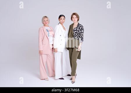 Drei verschiedene Frauen, darunter Asiatin und Europäerin, in eleganten Anzügen, die in voller Länge in einem hellen Studio stehen. Geeignet für Modeporträt, Power-Anzug und Stockfoto