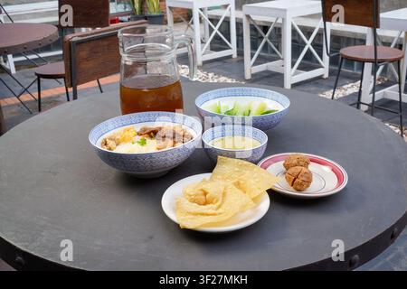 Asiatisches Nudelessen serviert mit knusprigen Wontons, Fleischbällchen, frischem Gemüse und einer Karaffe Eistee auf einem runden Cafétisch im Freien bei natürlichem Tageslicht Stockfoto