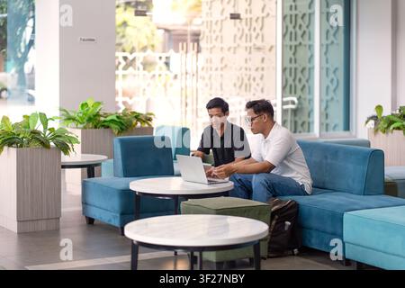 Asiatische indonesische Männer, die in einer modernen Bürolounge an einem Laptop zusammenarbeiten und Teamarbeit, Diskussion, Produktivität und Professionalität repräsentieren Stockfoto