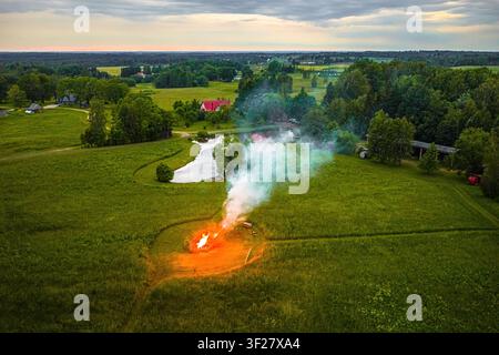 Luftaufnahme eines Lagerfeuers, das in einem grünen Feld brennt, mit aufsteigendem Rauch, einem kleinen Teich und einer ländlichen Landschaft im Hintergrund in Vidzeme, Lettland. Stockfoto