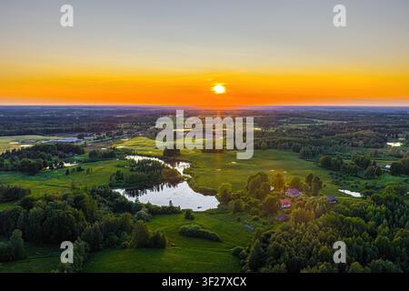 Luftbild einer ruhigen Landschaft mit einem spiegelnden See, umgeben von üppigen grünen Wäldern und Wiesen unter einem lebendigen goldenen Sonnenuntergang in Vidzeme, L Stockfoto