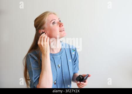Lächelnde Frau setzt echten kabellosen Ohrhörer ein, während sie die Ladestation hält, moderne Audiotechnologie und alltäglicher Lebensstil. Stockfoto
