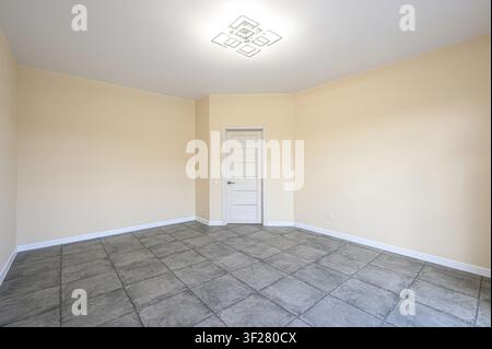 leerer Raum mit beigen Wänden, grauem Fliesenboden, einer weißen Tür und einer modernen Deckenleuchte Stockfoto