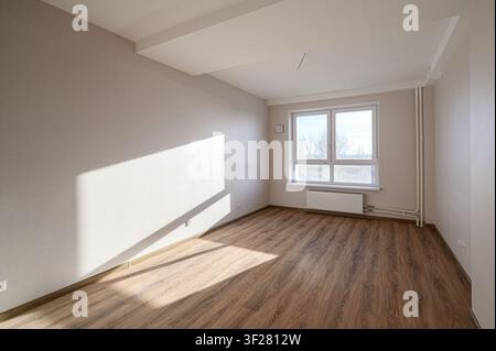 Ein leerer, heller Raum mit Holzfußboden, hellen Wänden und einem großen Fenster, das Sonnenlicht hereinlässt Stockfoto