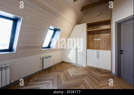 Dachzimmer mit schräger holzvertäfelter Decke und Wänden, Fenstern, Heizkörper, Fischgratboden und eingebautem Stauraum Stockfoto