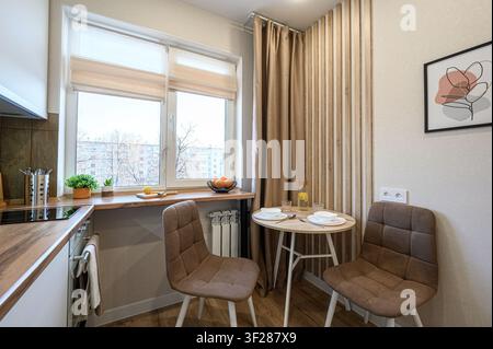 Gemütliche Küchenecke mit einem runden Tisch für zwei Personen, beleuchtet von einem Fenster. Neutrale Töne mit Holzakzenten schaffen eine warme, einladende Atmosphäre Stockfoto