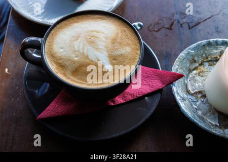 Überkopf-Nahaufnahme von Cappuccino mit cremigem Milchschaum, serviert in einer schwarzen Keramiktasse mit Untertasse auf einem hölzernen Cafétisch. Stockfoto