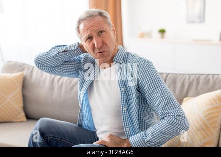 Älterer kaukasischer Mann, der Nackenschmerzen erlebt, während er zu Hause auf der Couch sitzt, Gesundheitskonzept Stockfoto
