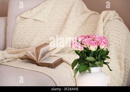 Offenes Buch auf einem gemütlichen Sofa mit einer weichen Überwurfdecke und einem Strauß rosa Rosen, das eine ruhige und einladende Wohnatmosphäre schafft Stockfoto
